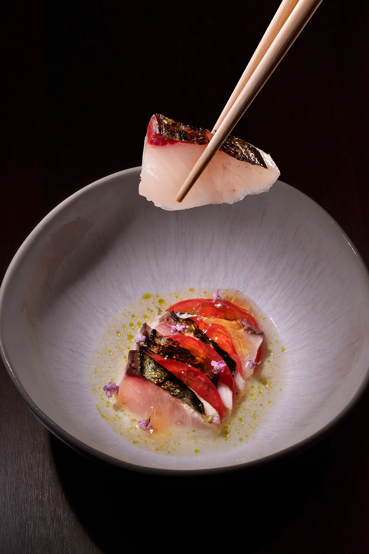 Sliced raw fish with tomato and herbs, with one piece held by chopsticks